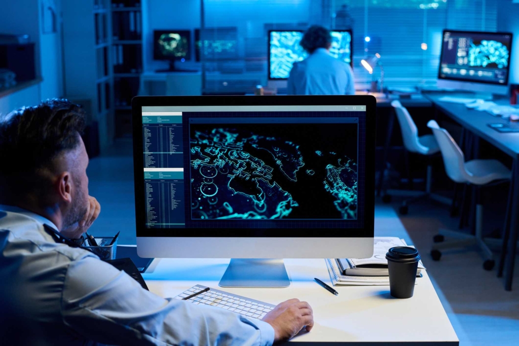 Patch Management for Compliance. A man sits at a desk in a dimly lit control room, facing a large monitor displaying a global cyber map with glowing data points. Multiple screens line the room, and another person works in the background. The environment feels tense and analytical, like a command center monitoring cyber threats in real time.